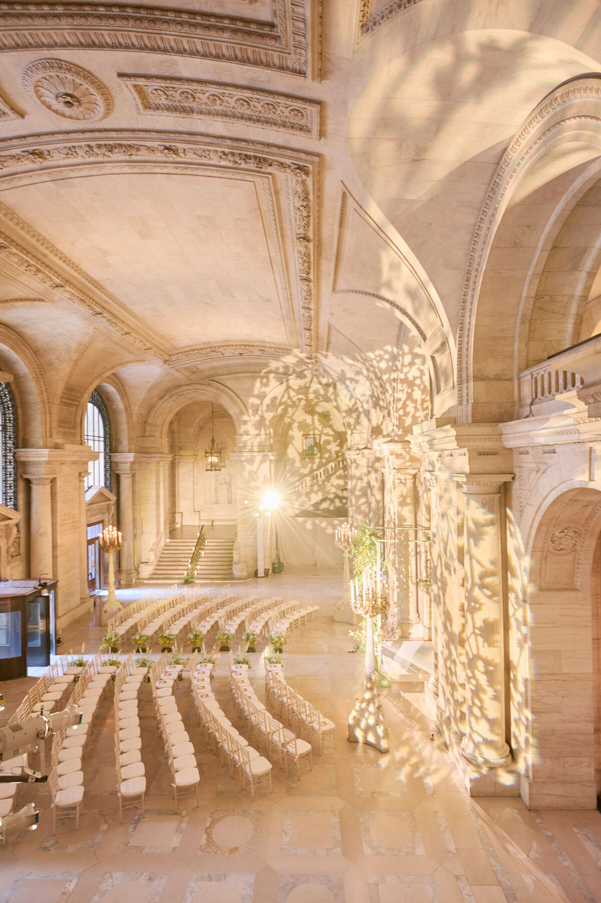 This stunning indoor venue, set for a wedding ceremony at the New York Public Library in New York City, features ornate architectural details, arched ceilings, and elaborate stonework. Rows of white chairs are artfully arranged in a semi-circle facing the altar, with warm, decorative lighting casting intricate patterns on the walls. The photo was captured by photographer Shawn Connell. The videographers for the event were Aaron Navak Films.