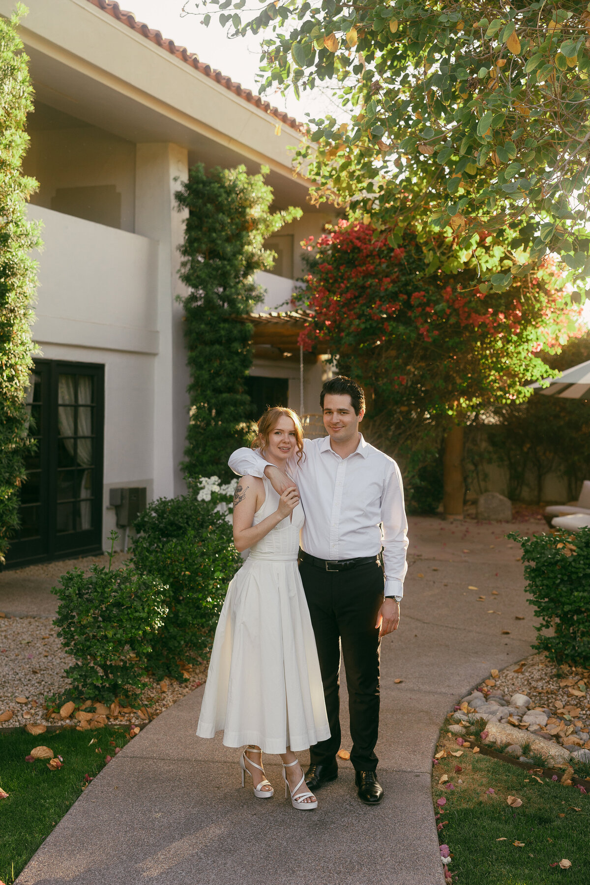 A man with dark hair stands next to a woman wearing a white dress with his arm over her shoulder while both look directly at the camera