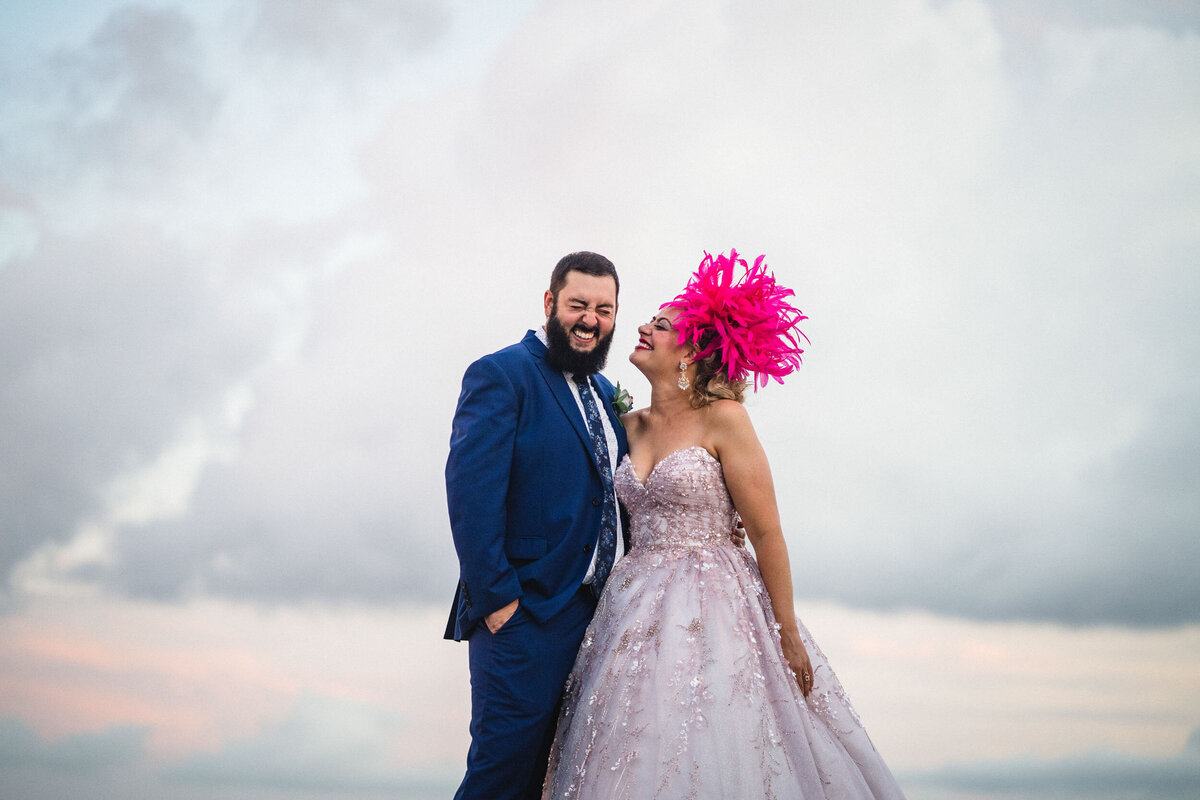 A bride and groom laugh heartily under a dramatic cloudy sky, capturing a moment of pure joy on their wedding day. The groom, in a sharp blue suit, embraces the bride who dazzles in a sparkling pink gown adorned with intricate beadwork. Her striking pink feather headpiece adds a whimsical flair to her ensemble, perfectly complementing her radiant smile and expressive makeup. This image showcases their vibrant personalities and the lively spirit of their celebration.