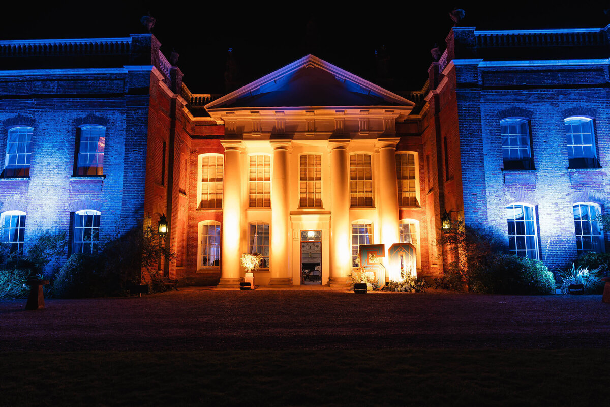 front facade of avington park lit up in gold and blue lighting at night for a 50th birthday party event planned  by westacott weddings and events