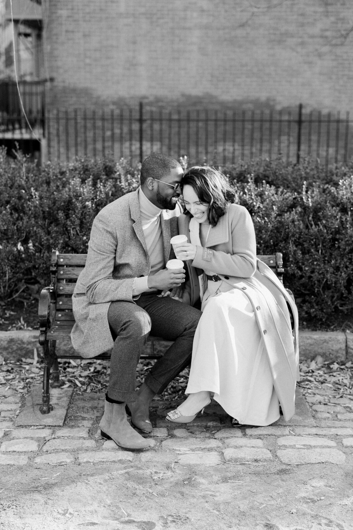 Couple sitting on a park bench close together as they both hold coffees.