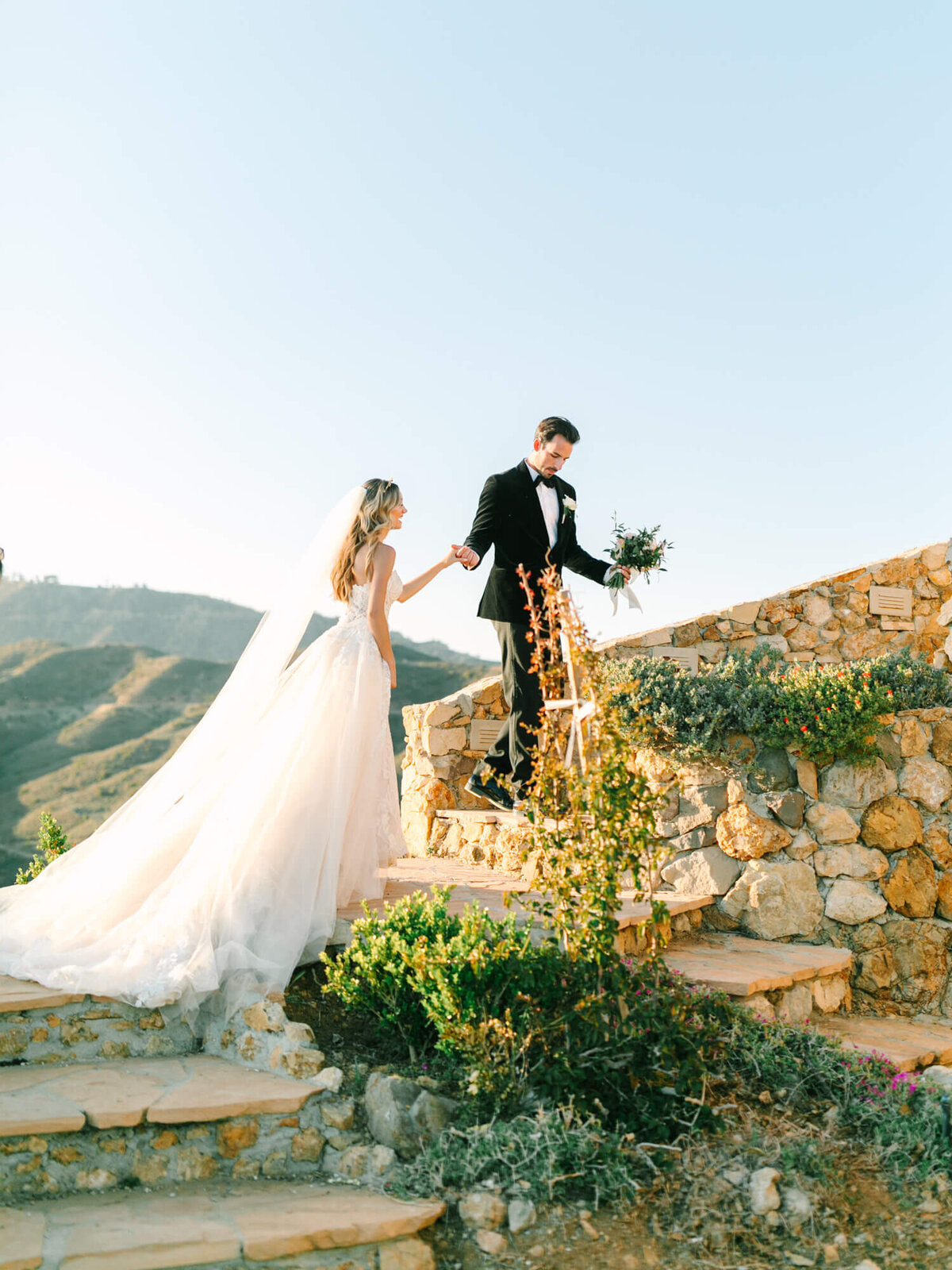 A bride in a flowing white gown and veil holds hands with a groom in a tuxedo on stone steps.