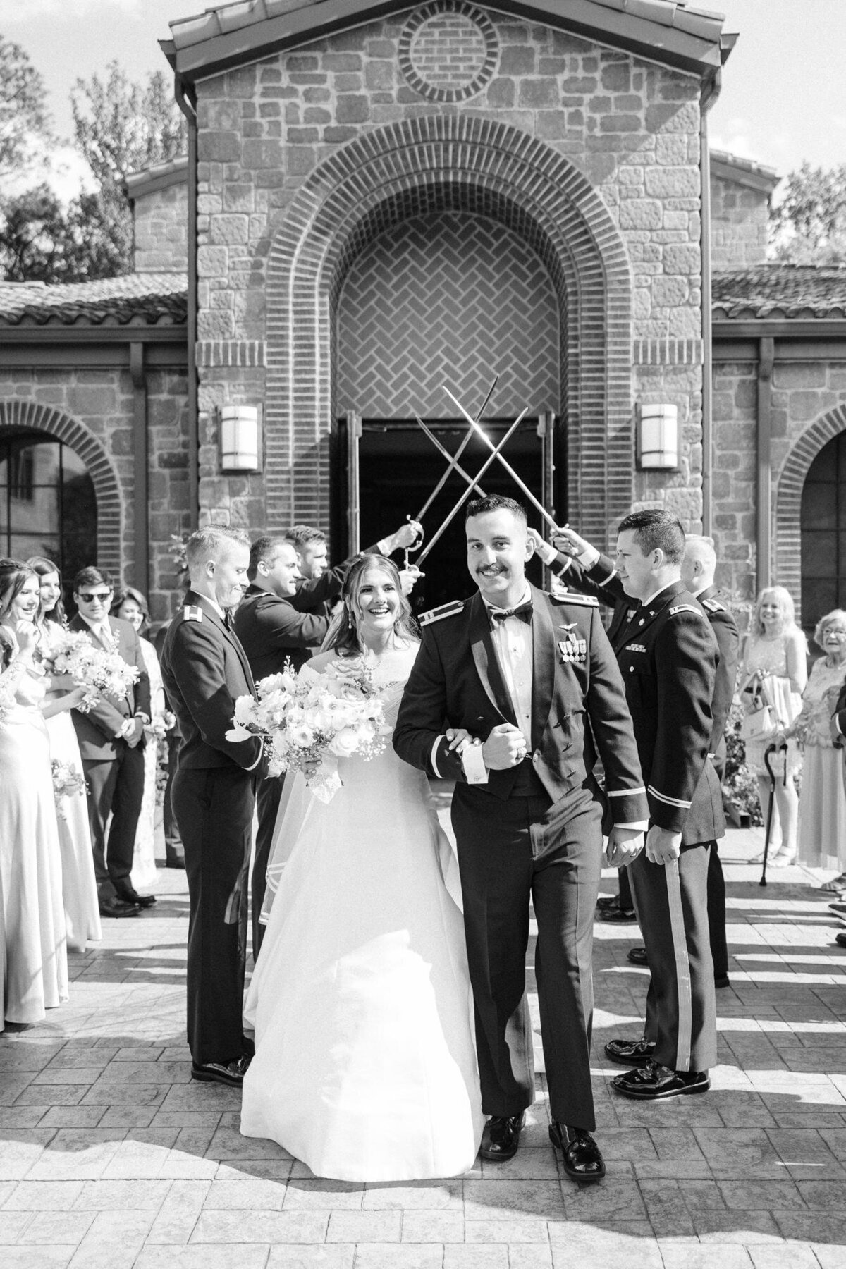 A newlywed couple exits a church while smiling as they pass under an arch of sabers held by uniformed military personnel.