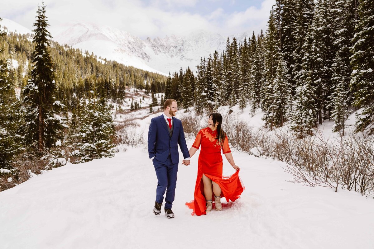 Couple runs through the snow hand in hand in the Colorado mountains while the bride wears a red dress during their Colorado elopement with Alpine Vows
