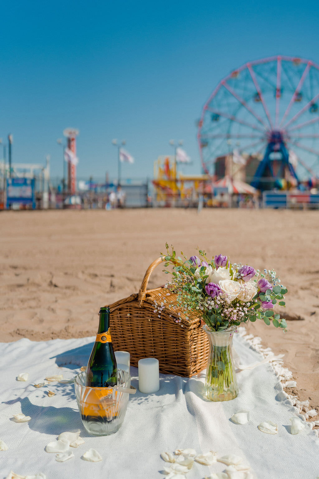 flowers, a champagne bottle chilling, candles, and a picnic basket laid out on a blanket in the sand