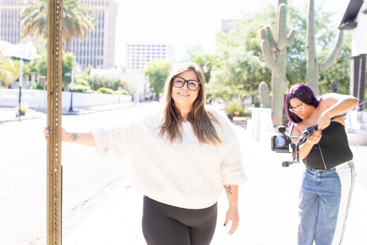 Smiling woman in a fluffy white sweater posing outdoors on a sunny Tucson street, holding onto a signpost, photographed by Vyrl Photo, showcasing Tucson brand photography.