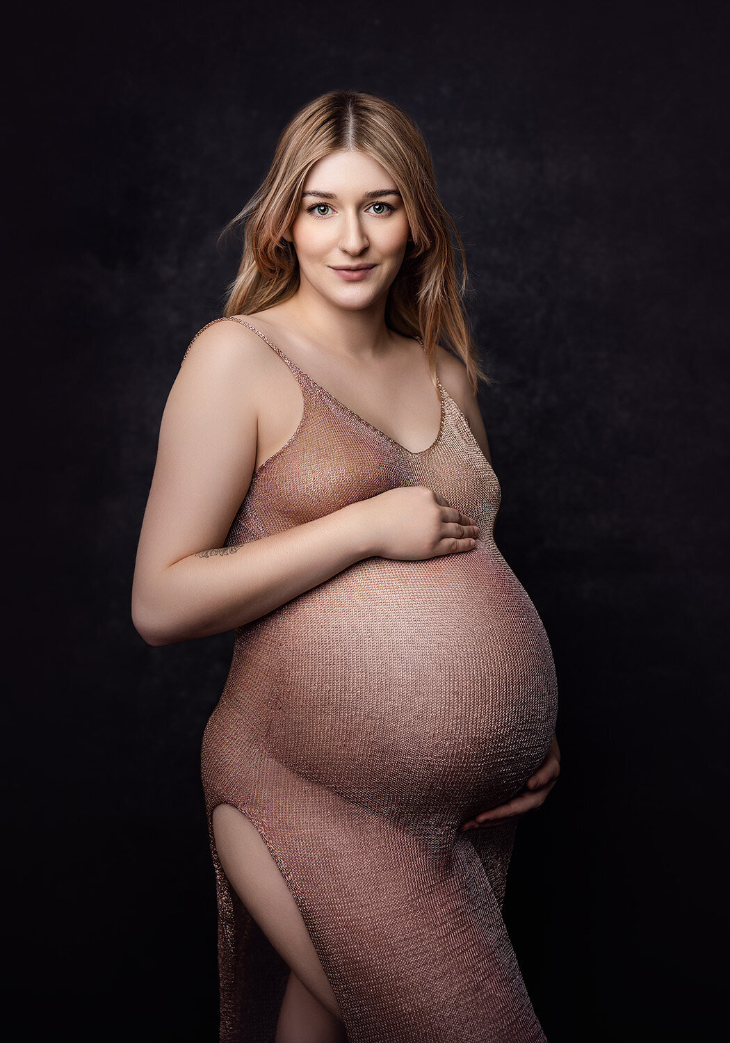 Image of a pregnant woman cradling her bump in a sheer pink dress. She is standing on a black background with her hair blowing in the wind. Taken by Norwich maternity photographer Claire Howes.