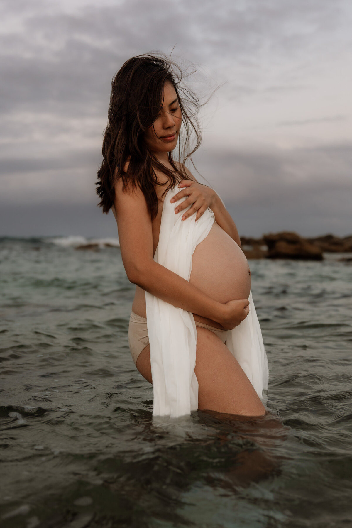 Pregnant woman standing in ocean water with flowing white fabric during a serene beach maternity session in Sydney.