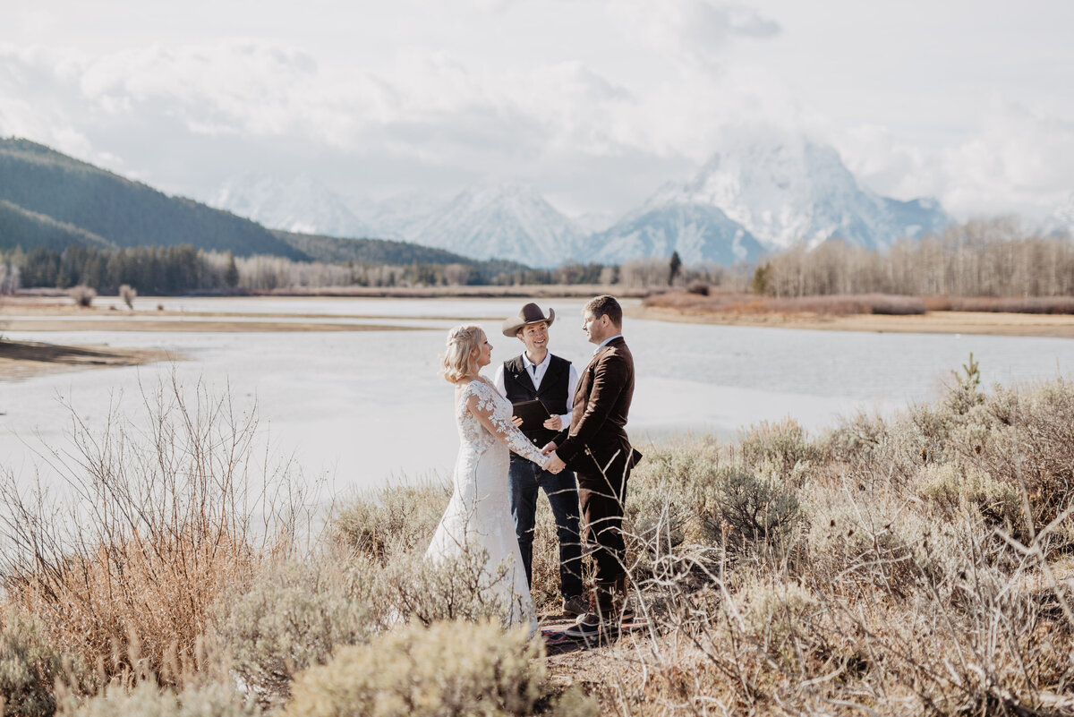 outdoor winter ceremony with bride and groom holding hands while they say their vows in front of a frozen river captured by jackson hole wedding photographers