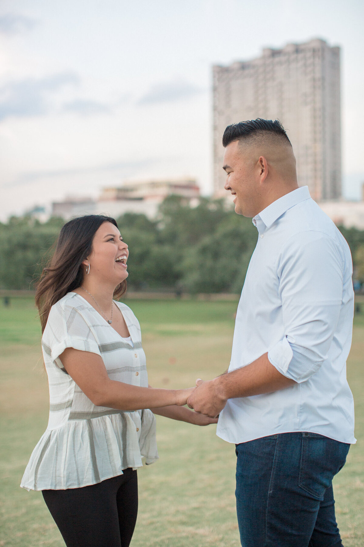 A woman laughs joyfully while holding her betrothed's hands in front of a city skyline