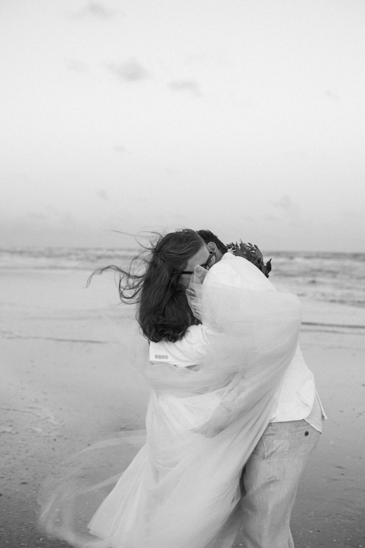 A black and white photo of a bride and groom deeply embracing on the beach after their wedding ceremony at Crystal Beach, Texas. The bride is on the left and is wearing a white dress with a long, flowing veil that wraps around the front of both of them. The groom is on the right and is wearing a white dress shirt with khaki pants.