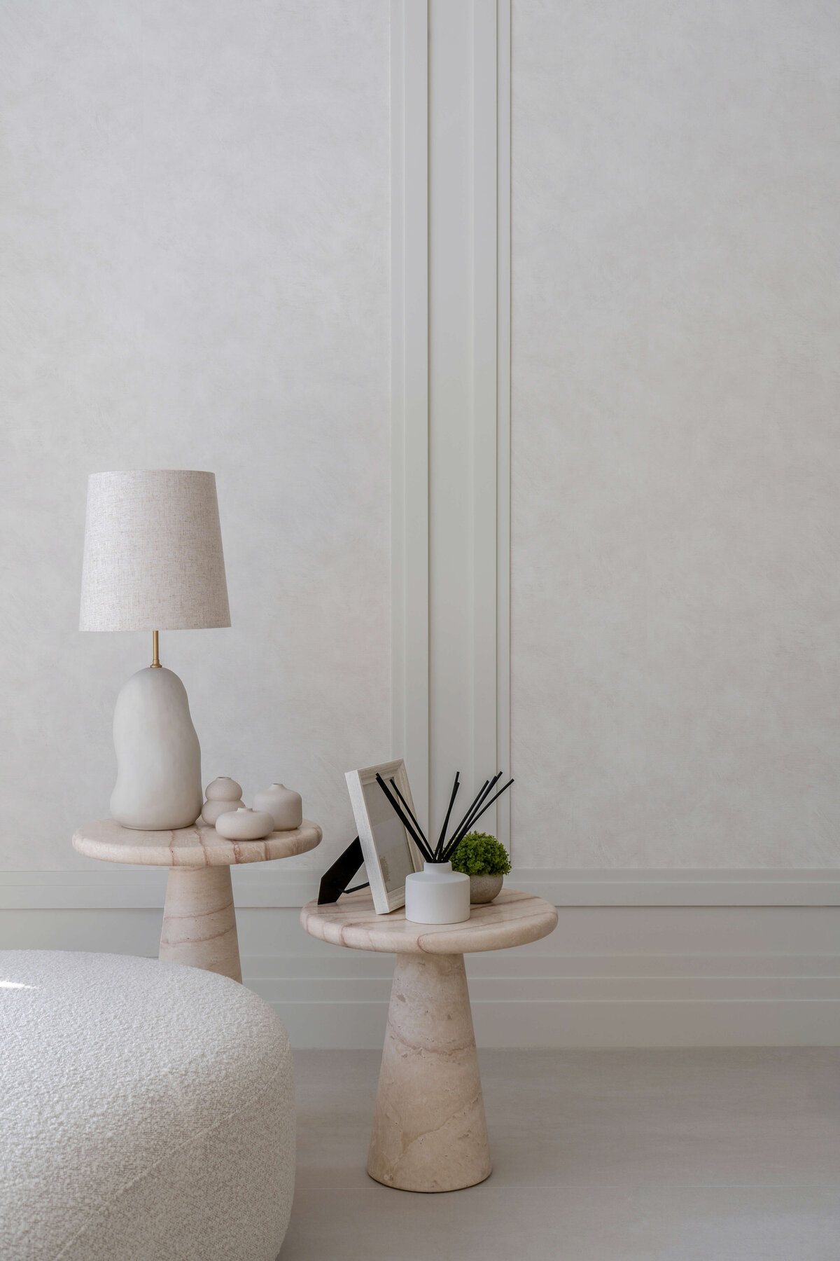 Two small, light marble tables. The left table has a lamp and decor. The right table has a photo frame, a reed diffuser, and plant.