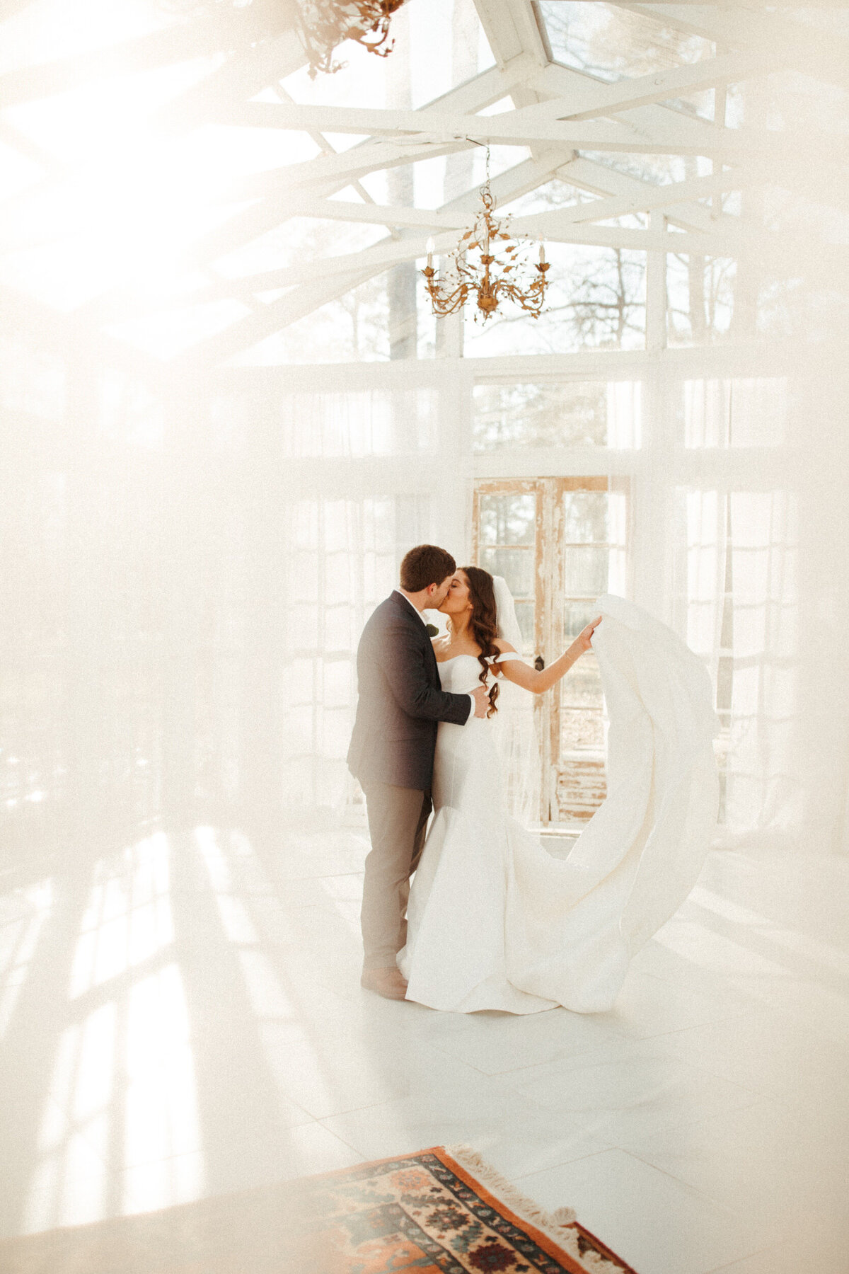 Bride and groom standing inside atelier while kissing as the sunlight pours in around them