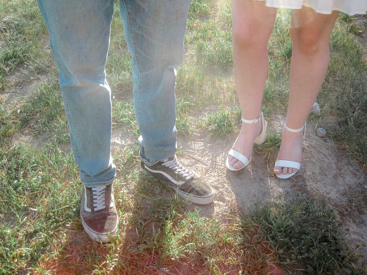 Close-up of a couple’s feet standing on grass, with the man wearing sneakers and jeans, and the woman in white high-heeled sandals and a light dress, emphasizing their contrasting casual and formal footwear.