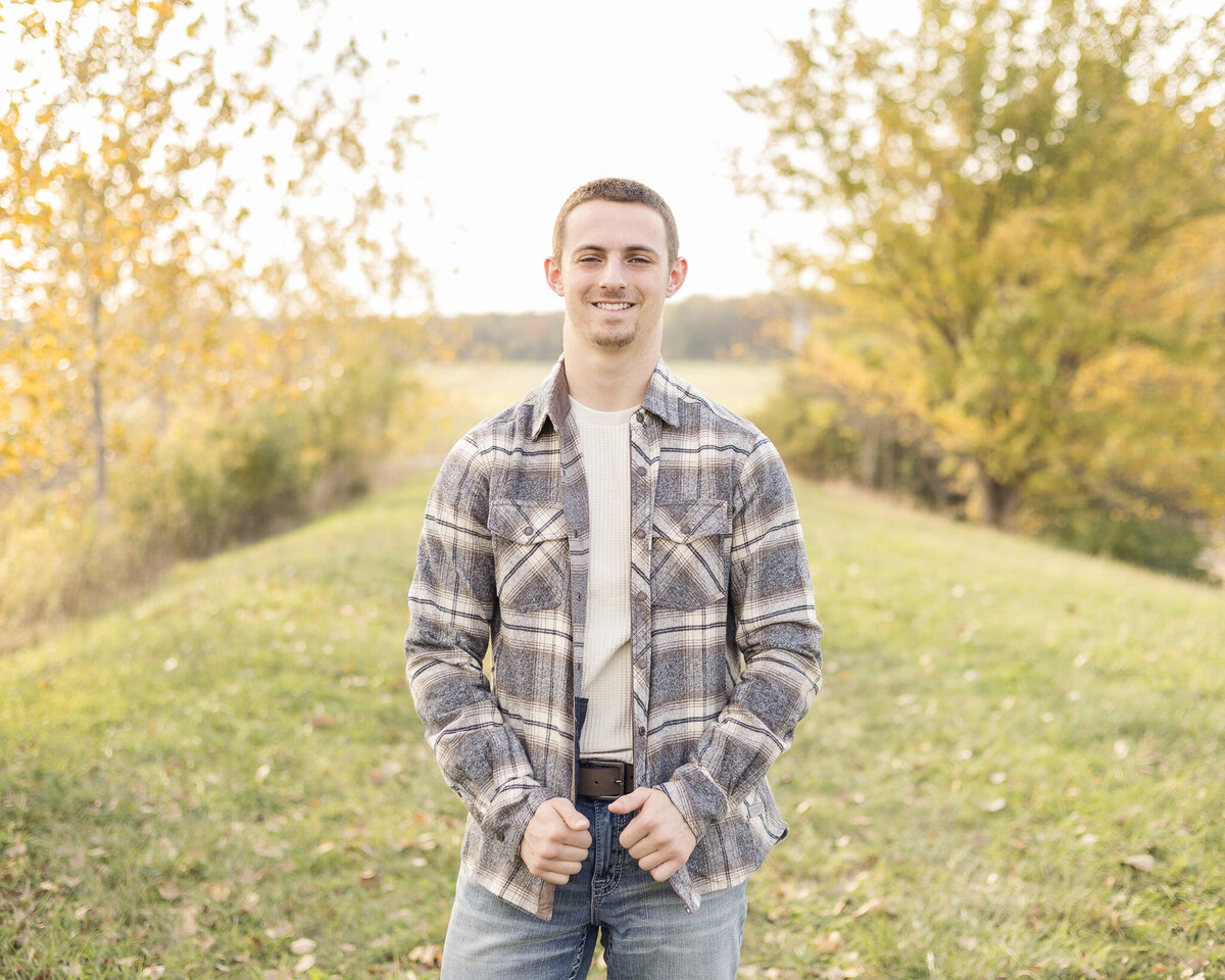 A high school senior male, standing in the grass, with trees in the background, wearing a flannel jacket, light tan long sleeve shirt, blue jeans, short brown hair, and smiling at the camera