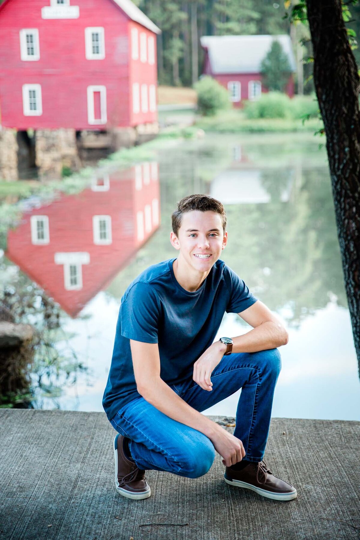 Handsome senior squatting in front of a  pond with a reflection of a red barn in the water.