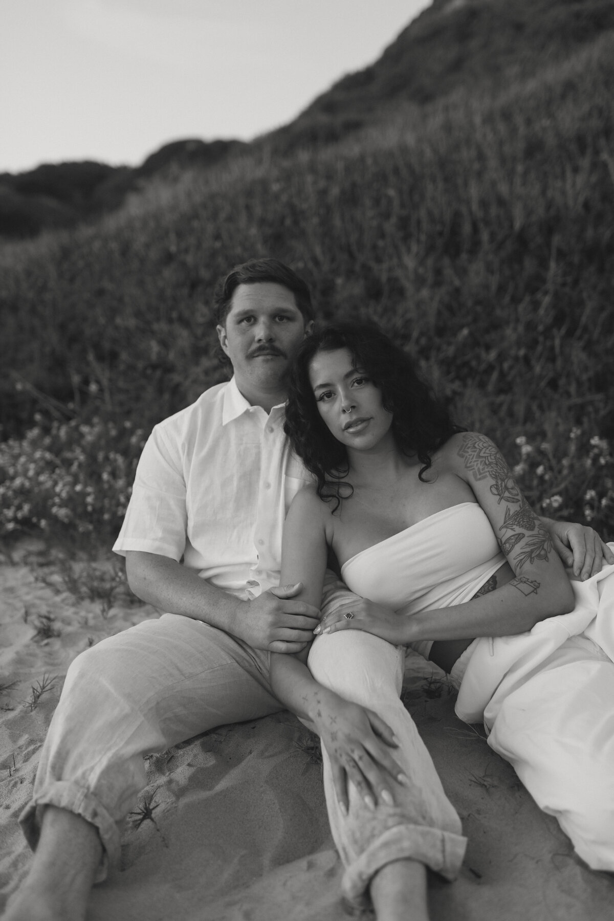 engagement photos on the beach sitting next to each other shes leaning into his side looking seriously at the camera