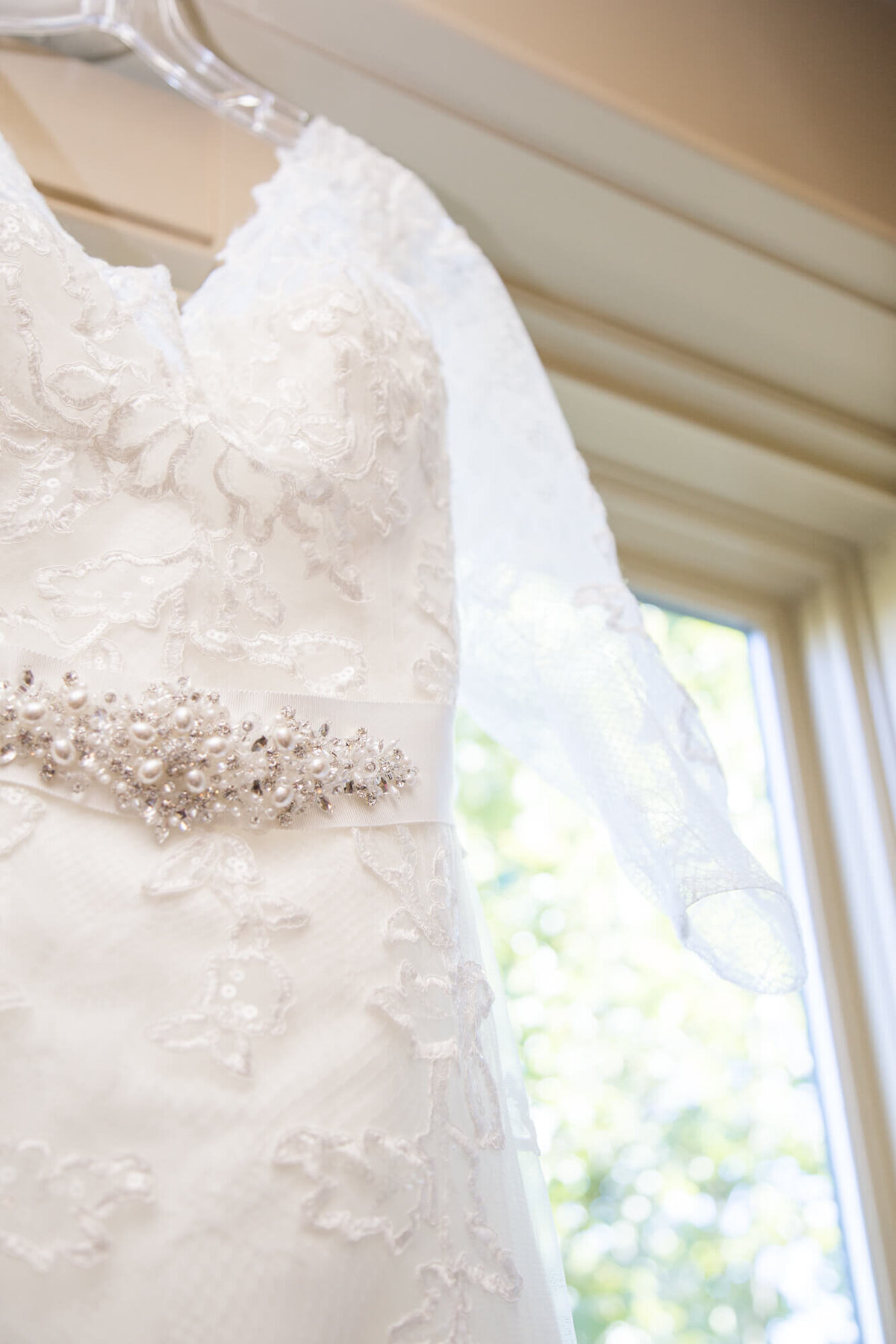 calssic wedding detail of lace dress with beaded belt hanging by a window