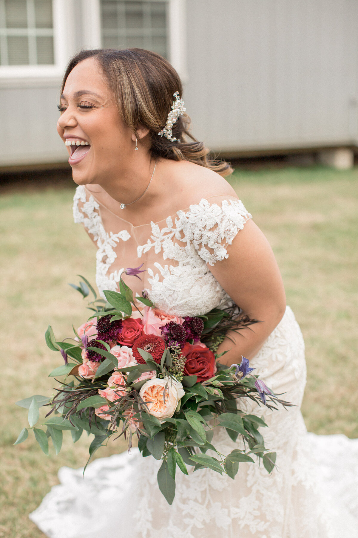 A bride in a lacy wedding dress laughs, holding a bouquet of red and pink roses.