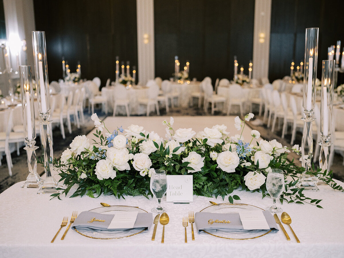 A classic Calgary wedding at the Fairmont Palliser boasts a head table adorned with white flowers, greenery, and place settings for two. Tall candles and elegantly set tables are visible in the background, creating a timeless ambiance.