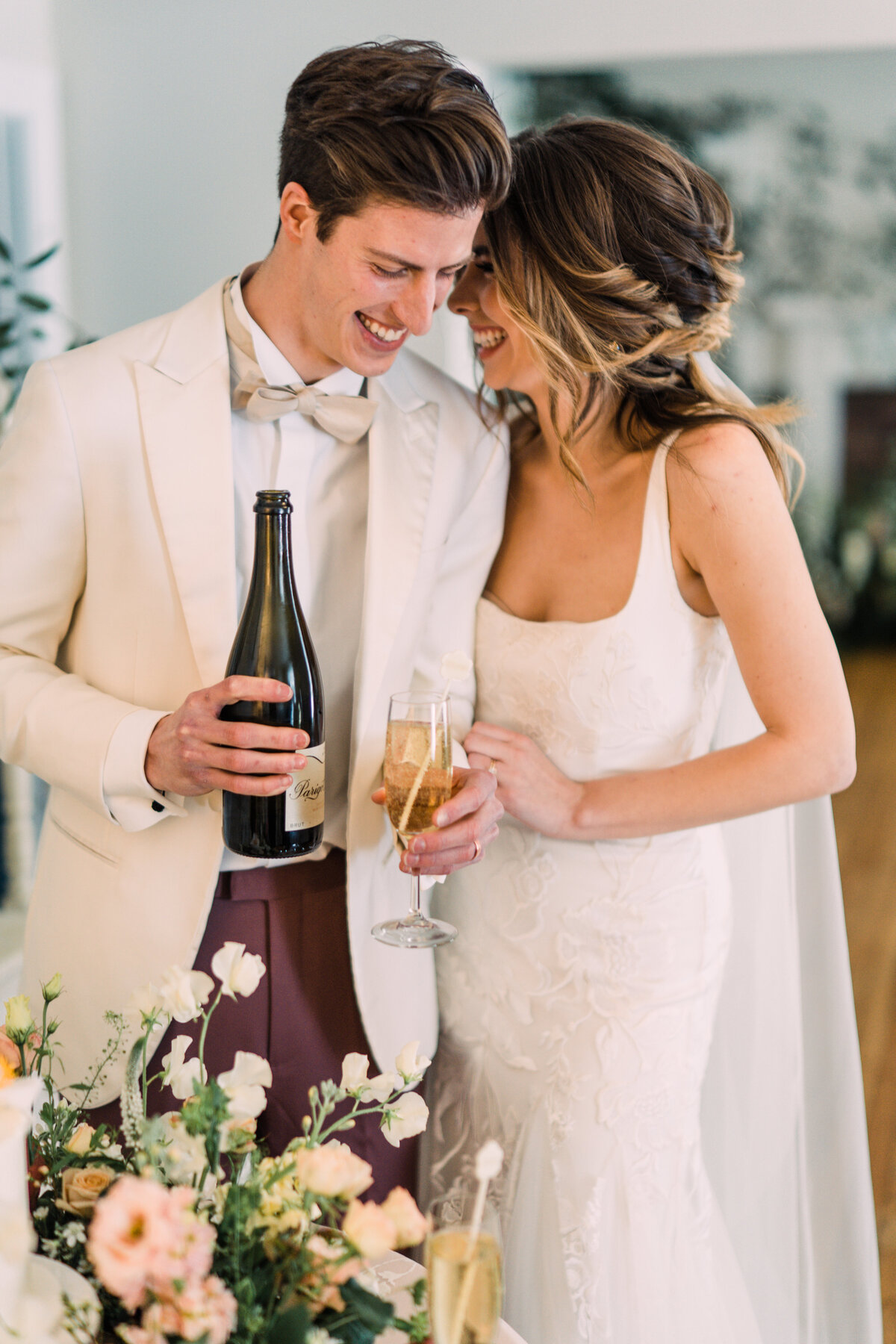 Bride holds groom's arm and laughs while he smiles and looks at a bottle and glass of champagne with flowers in foreground