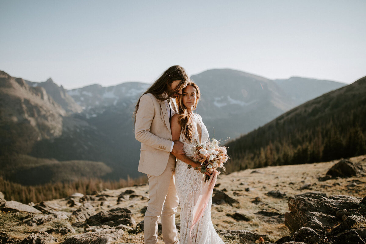 Bride and groom wearing tan wedding suit and white wedding gown hold each other with bouquet of flowers in the mountains.