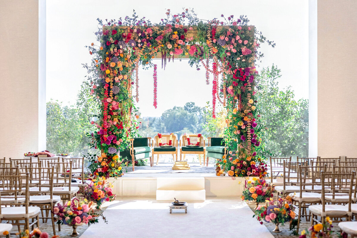 A beautifully decorated wedding altar with vibrant floral arrangements in shades of pink, orange, and purple. Green chairs are placed beneath the arch, and rows of wooden chairs line the aisle leading to the altar.