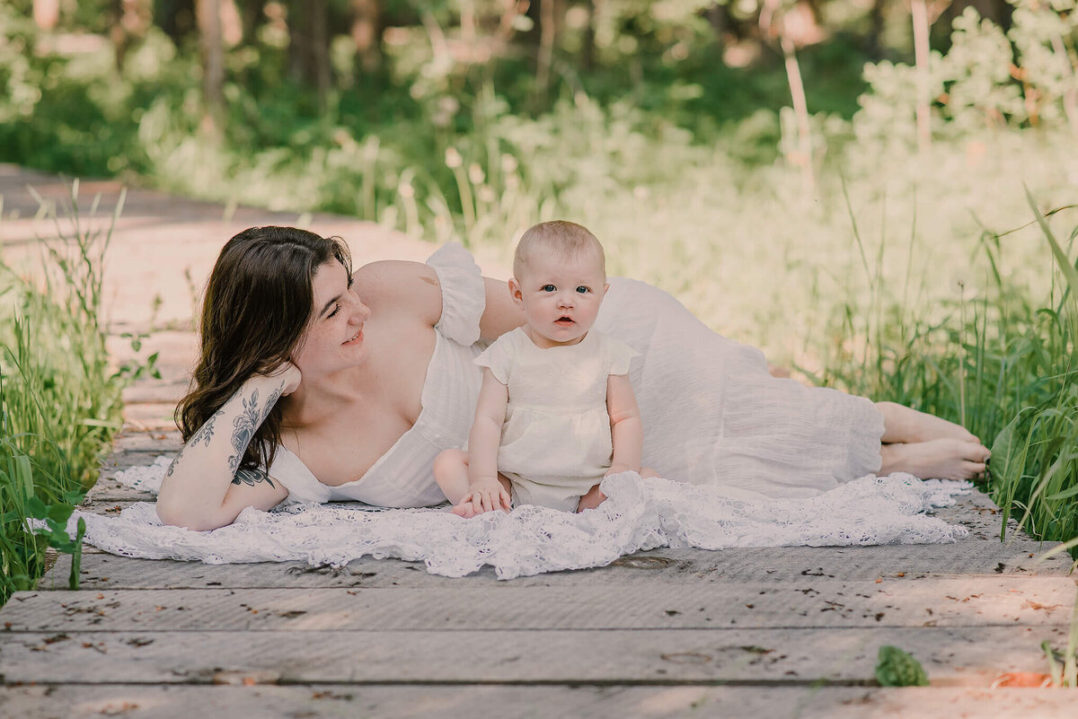 a Mother and daughter dressed in white lay on a wooden bridge. Mother smiles at duaghter while daughter looks at the camera.  Captured by Brittany Danielle