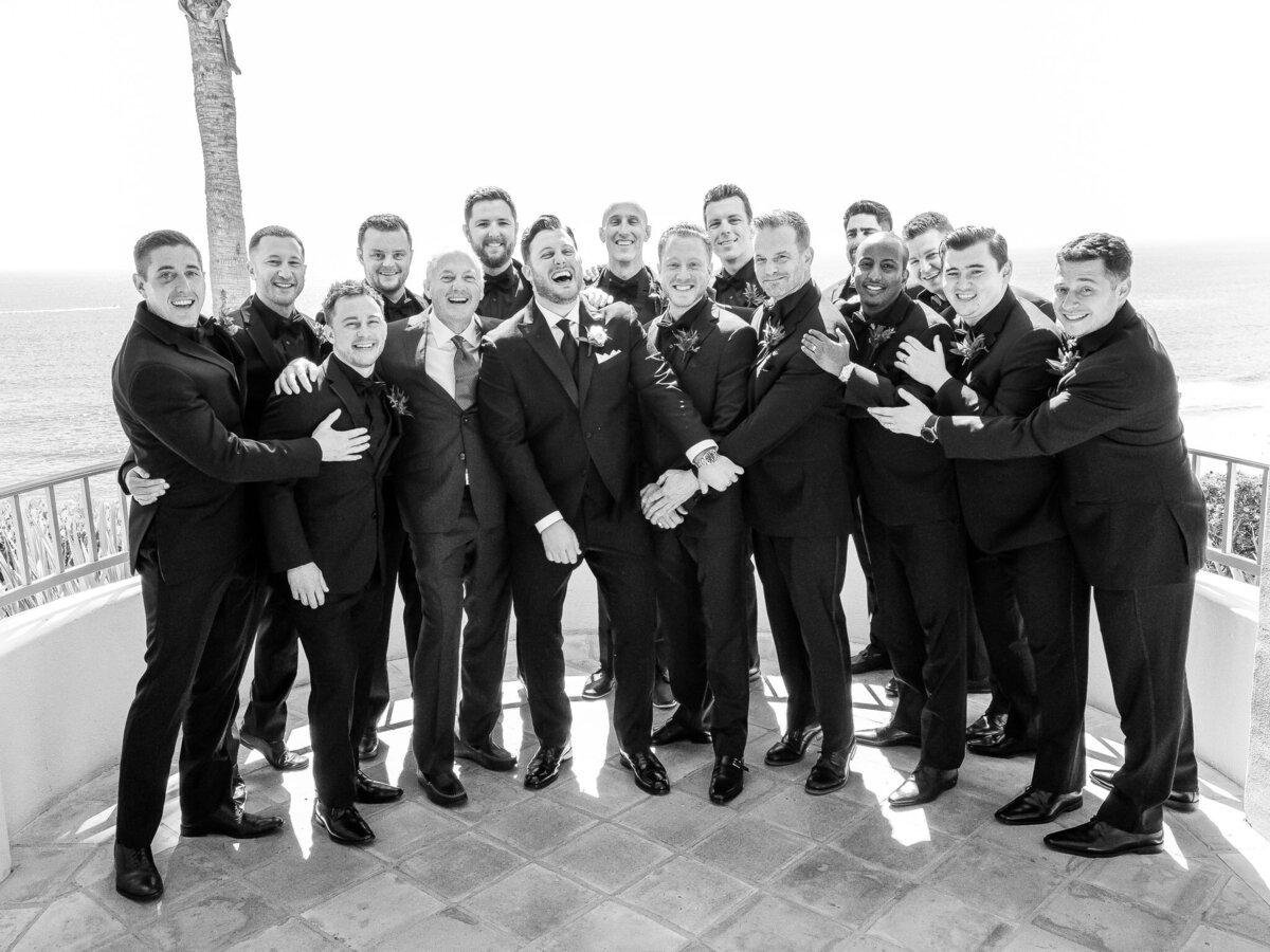 The groom and his groomsmen, dressed in matching black suits, share a joyful moment on a balcony overlooking the ocean at Ritz-Carlton Laguna Niguel