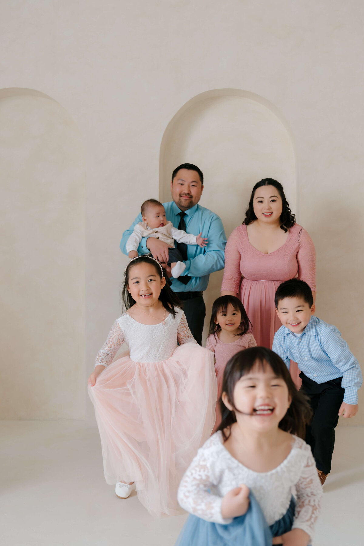 Children laughing and running toward the camera while parents hold baby in the background, all smiling in a soft-lit studio.