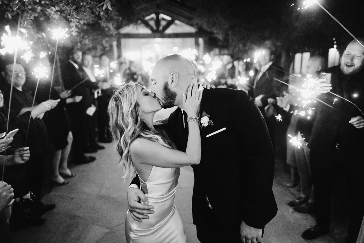 Romantic sparkler exit at Roblar Winery in Santa Ynez Valley. Black and white documentary photography capturing newlyweds' passionate kiss amidst sparkler send-off. Bride in sleek silk slip dress and groom in classic black tuxedo. Joyful wine country celebration under the rustic wooden pergola, surrounded by vineyard ambiance. Timeless Santa Barbara wine country wedding moment with ambient sparkler lighting and candid guest reactions. Luxury Santa Ynez Valley wedding photography.