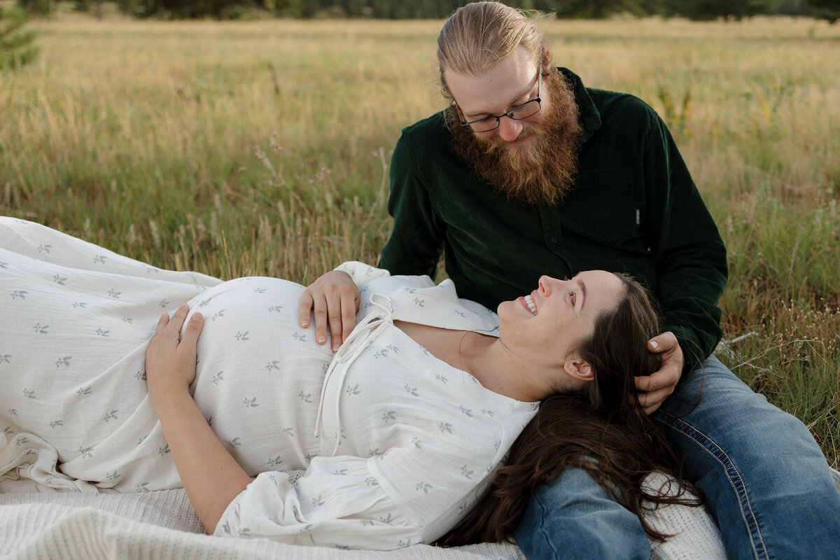 Expectant parents laying on white blanket in a field looking at each other and smiling in Boulder Colorado