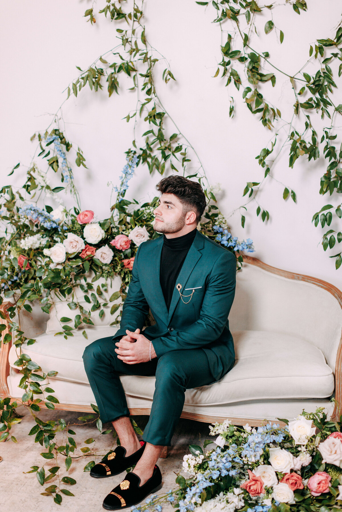 groom wearing emerald colored suit on a love seat surrounded by florals