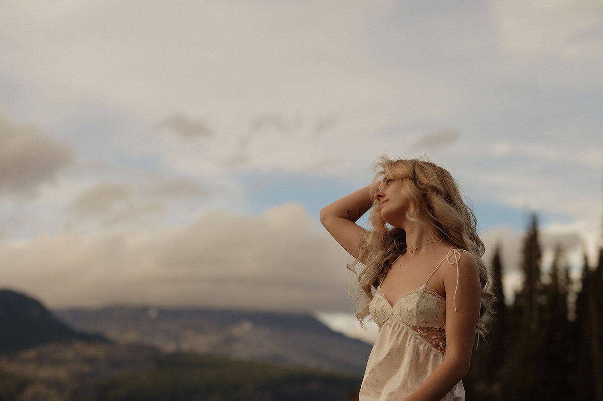 woman touches her hair while she stands in front of a cloudy sky backdrop at Sparks Lake