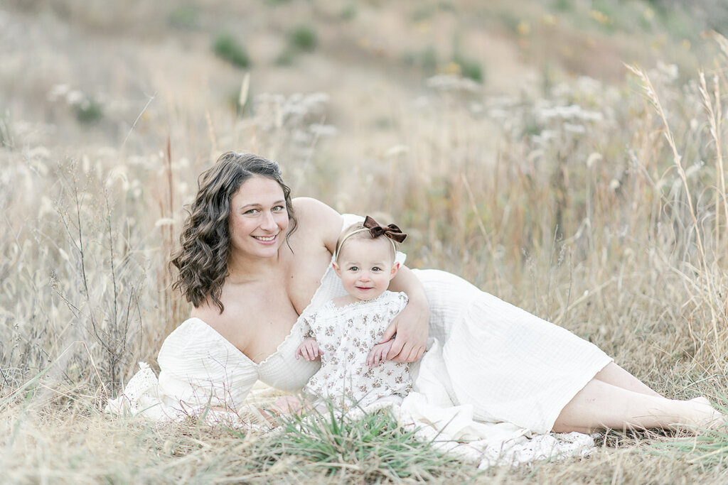 Mother lays on the ground smiling with her baby sitting in a field