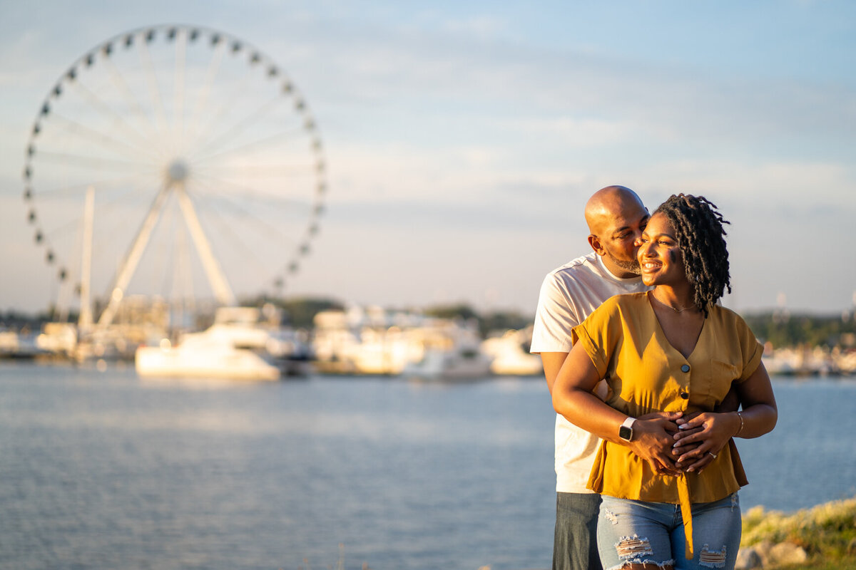 An African-American couple smile at each other with the National Harbor ferris wheel behind them.