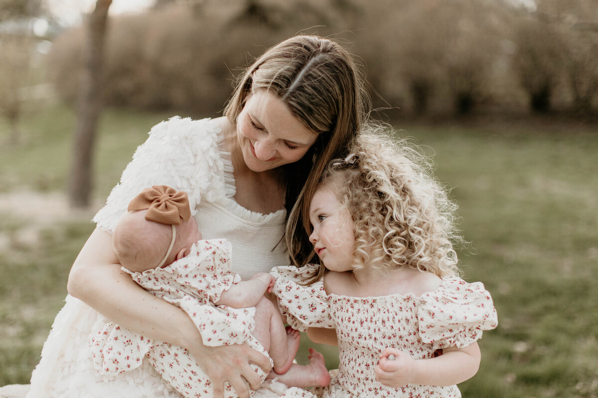 Mom with both girls sitting on a stone while the toddler girl holds baby's hand.