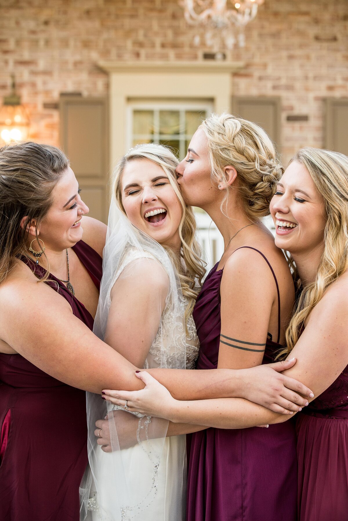 a bride is hugged tightly by her three bridesmaids who wear maroon dresses