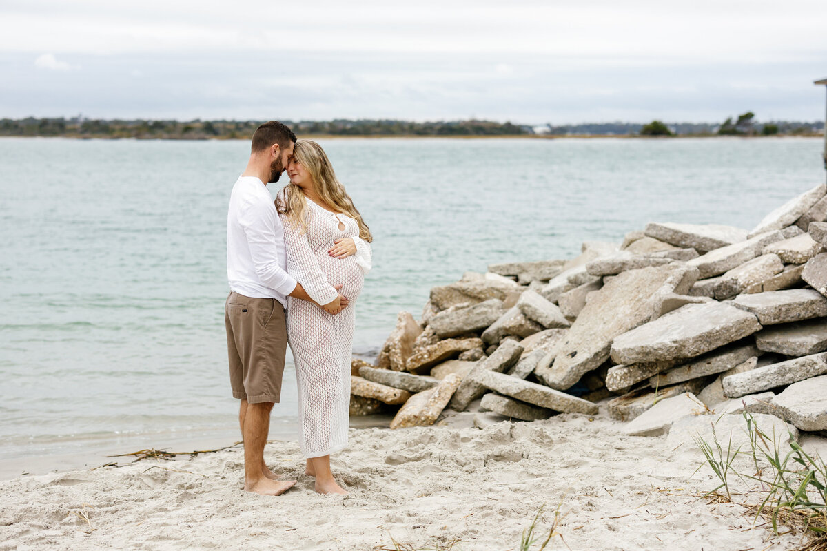 Husband hugging pregnant wife from behind on Wilmington beach, both touching baby bump
