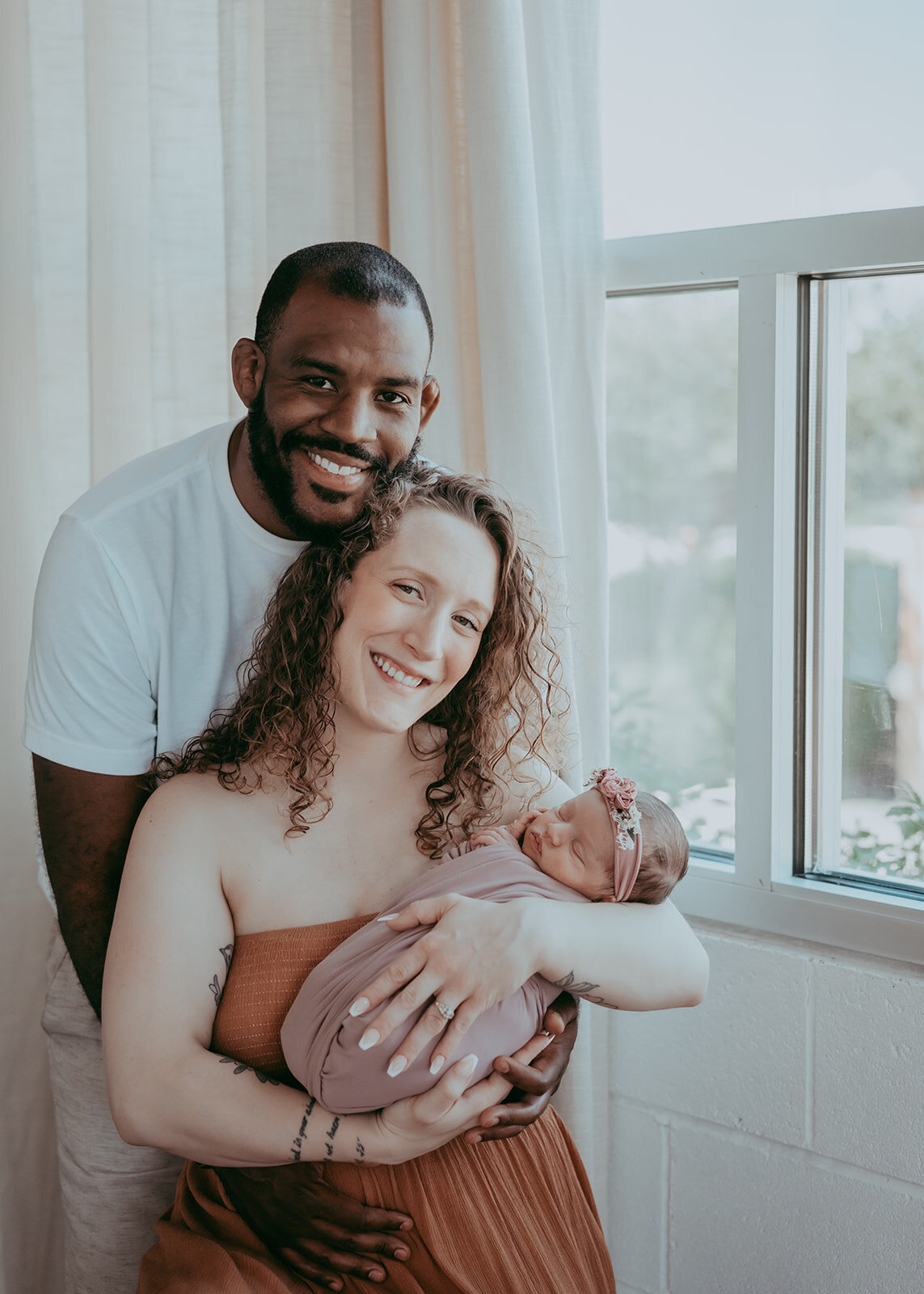 newborn couple smiles at camera while holding their swaddled baby girl
