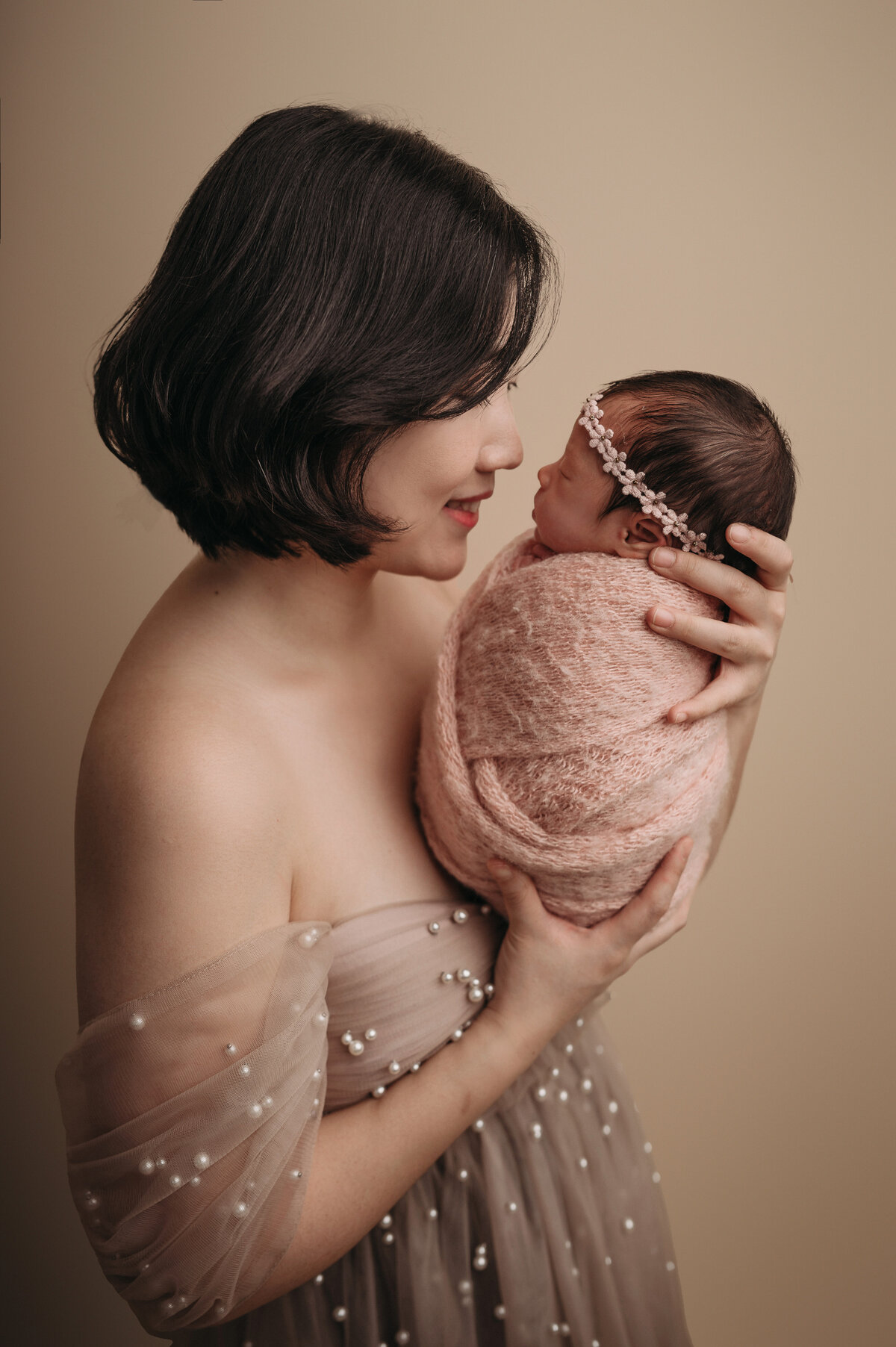 new mom with short brown hair wearing a cream pearl gown  turned to her side holding and looking  her newborn baby girl swaddled in a pink  blanket and wearing a floral pink headband in  front of cream backdrop