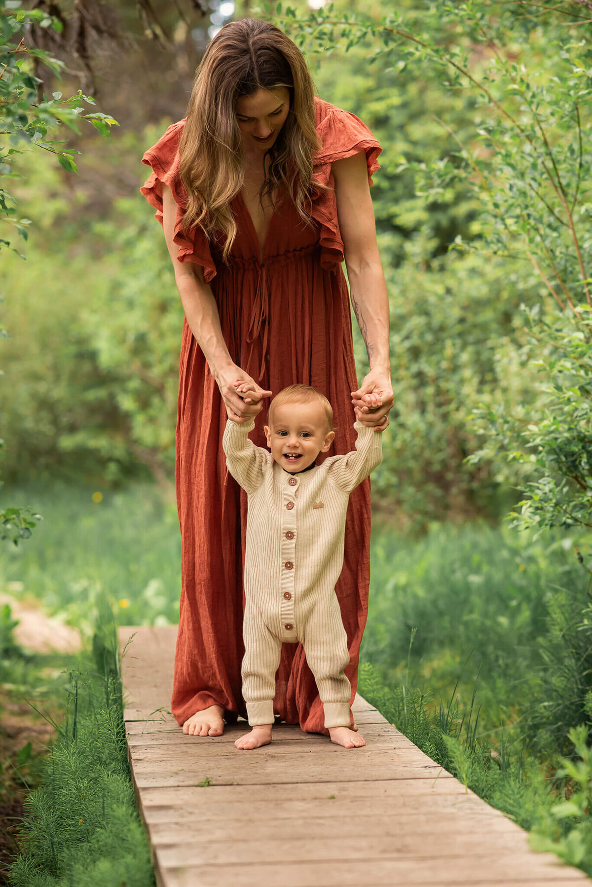 A mom is standing on a little wooden bridge over a creek. She bends over and holds the hands of her infant son while he tries to walk accross. He smiles. Portrait by Brittany Danielle Photography