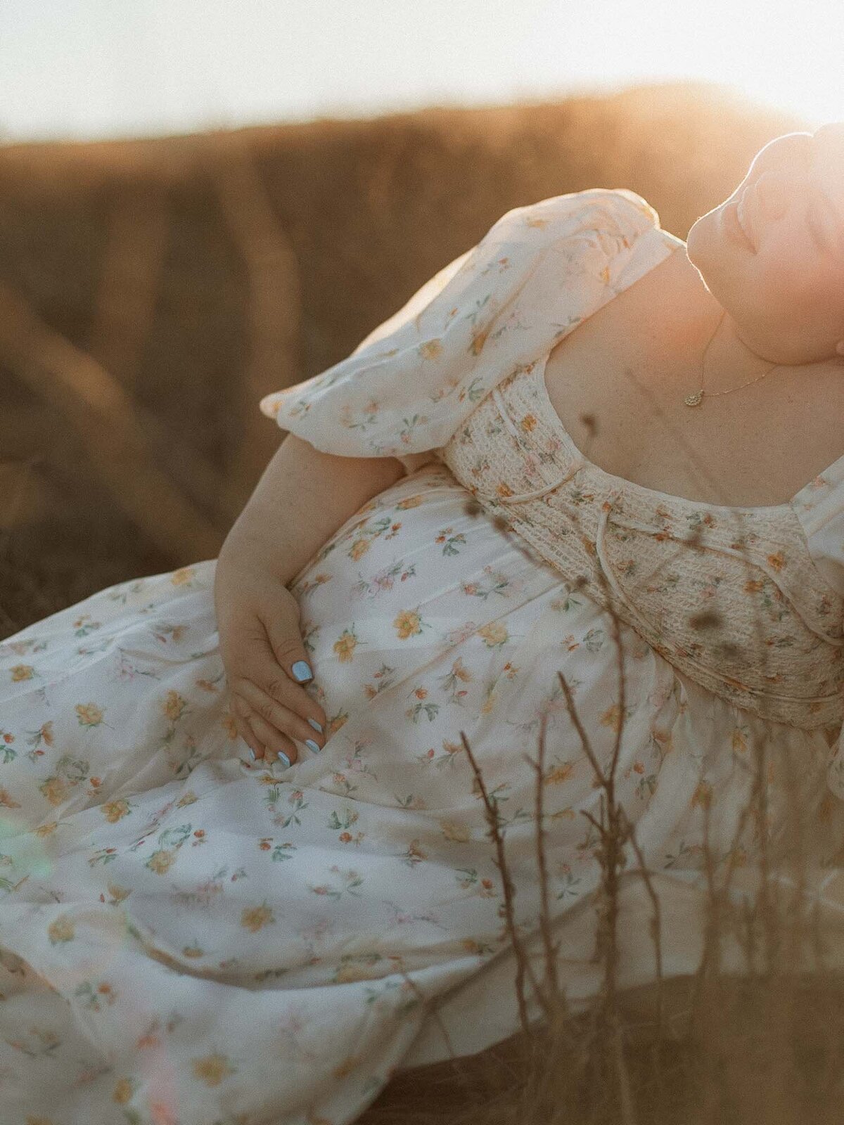 Expectant mother sitting in a golden field at sunset, holding her bare belly with both hands.