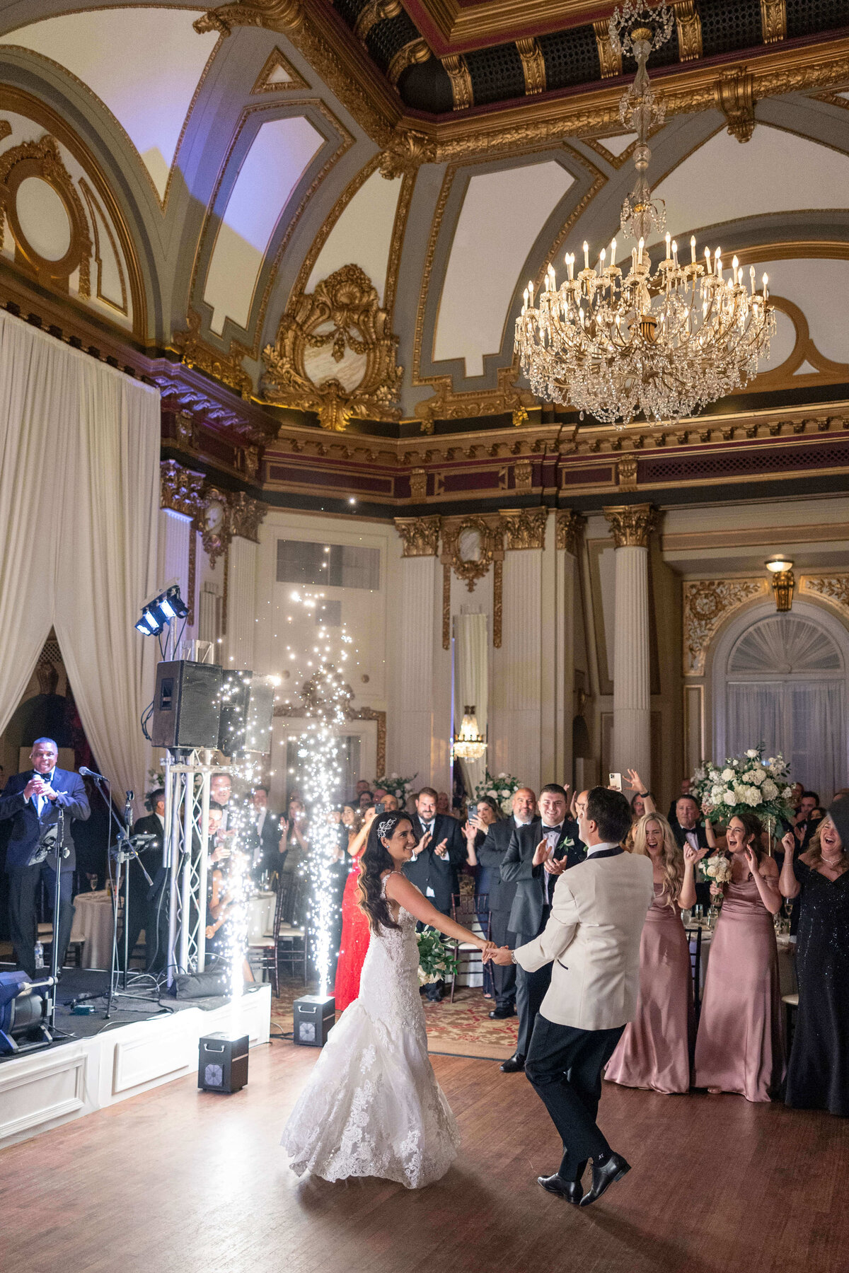 A couple dances joyfully in the center of an elegant, ornate ballroom with a chandelier above. Friends and family are cheering them on, and a band plays in the background. Sparkling fireworks light up the scene.