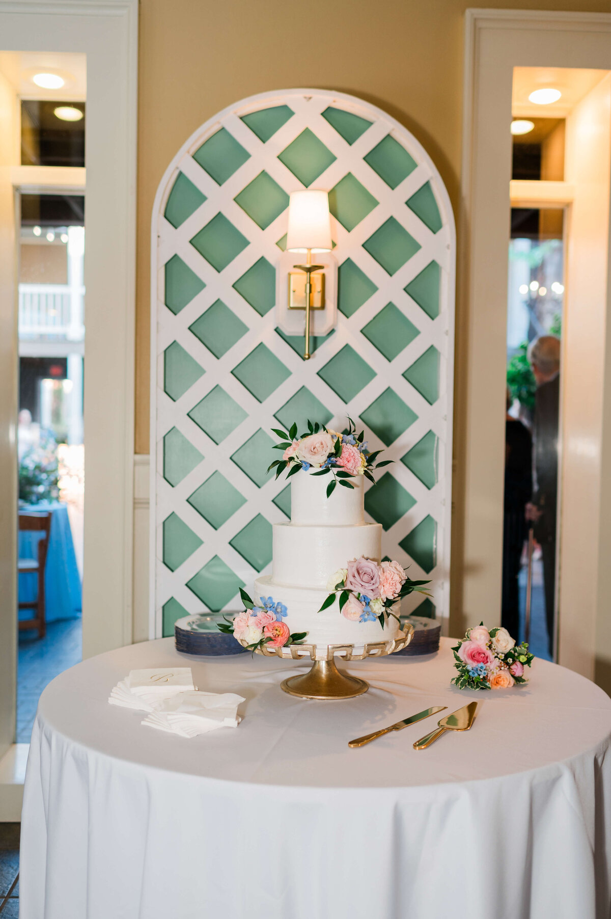 A white two-tiered wedding cake with pastel flowers sits on a gold stand atop a round table with a white tablecloth. Floral arrangements and cutlery are arranged on the table, with a decorative white lattice backdrop behind.