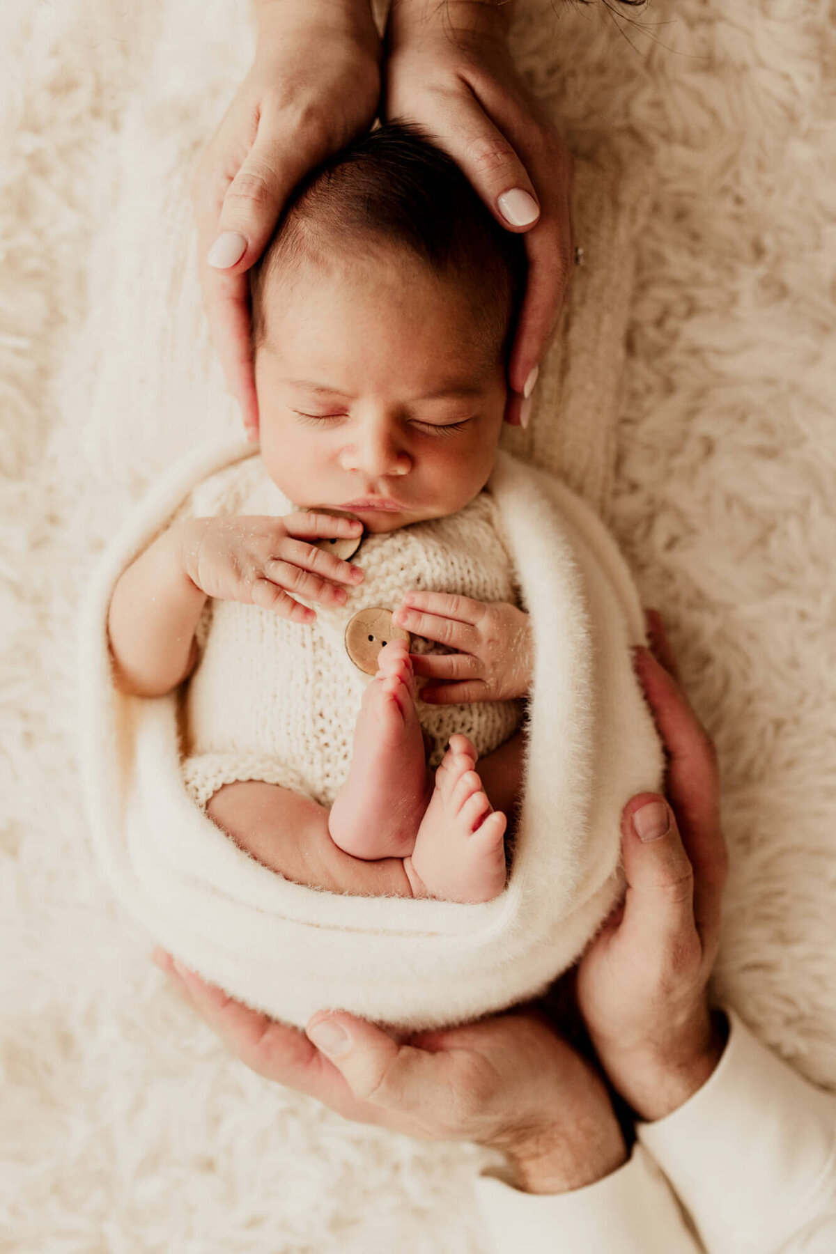 Baby boy wrapped in a cream wrap with his mother and father's hands placed on him.