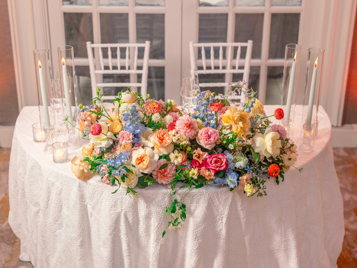 A beautifully decorated table with a white tablecloth, adorned with an arrangement of colorful flowers including pink, yellow, and orange blooms. Clear glass candles in tall holders are placed around the floral centerpiece, creating an elegant setting.