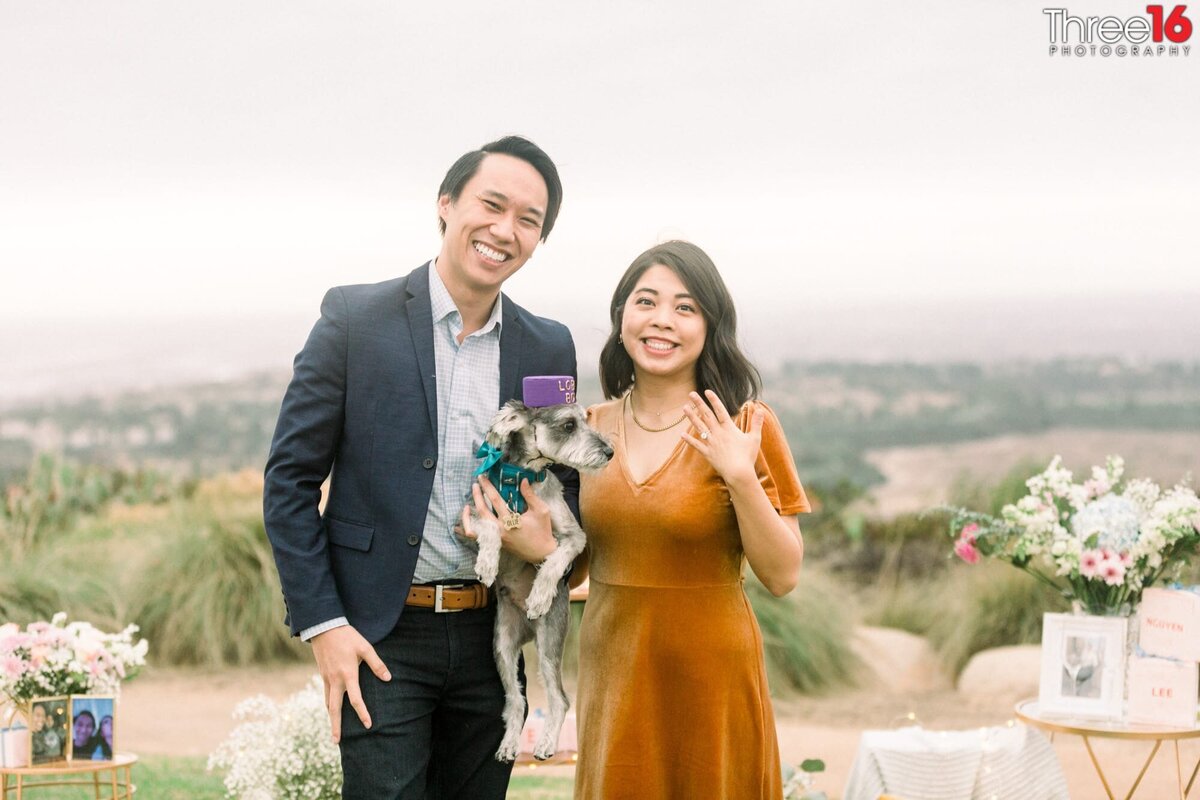 Newly engaged couple and their dog smile for the photographer and shows off the ring after a marriage proposal