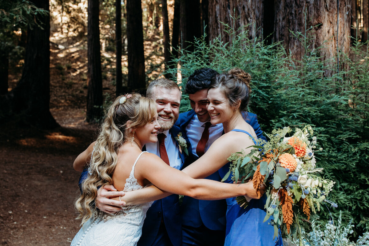 A joyful bride and groom embracing friends in a forest, with a large, colorful bouquet in the foreground.