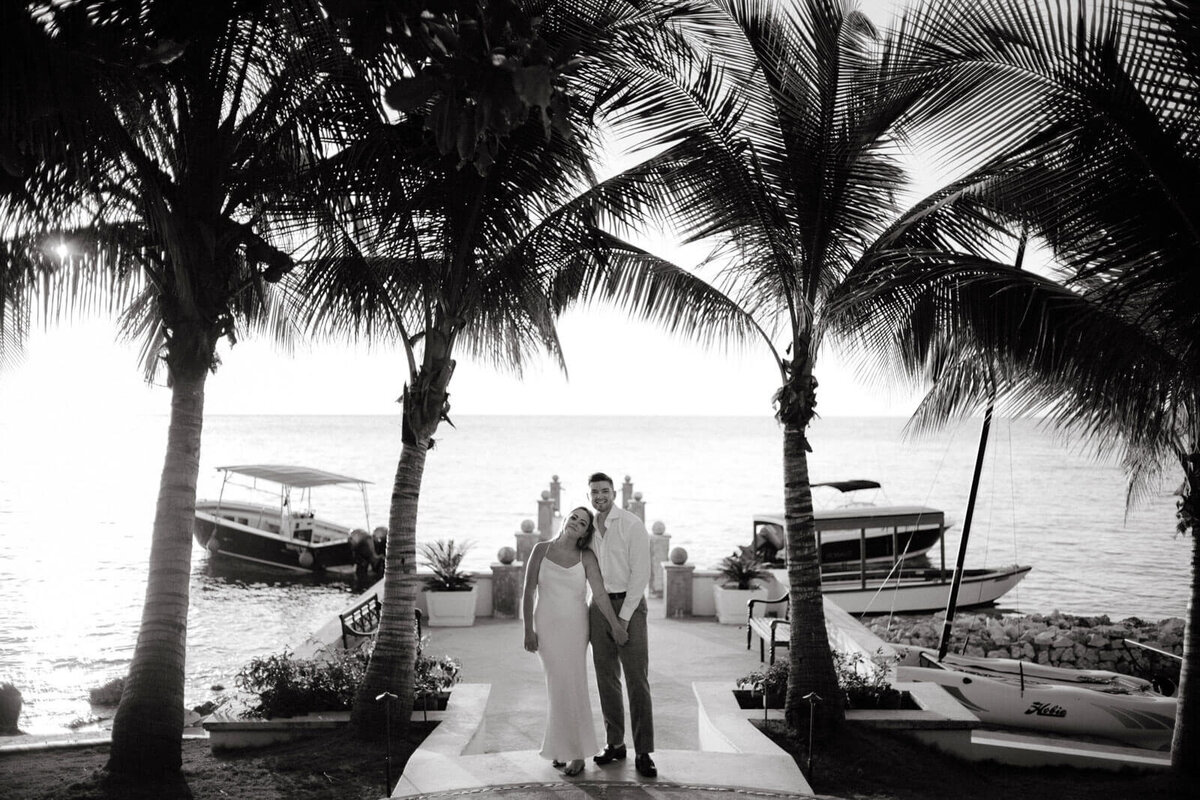 Black and white photo of the engaged couple standing in front of the ocean, with small boats and coconut trees in Jamaica.
